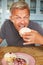 Mature Man Sitting At Table Eating Sugary Donut