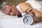 Mature man sleeping in bed with alarm clock in foreground at bedroom. Exhausted man being awakened by an alarm clock in his