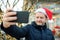 Mature man wearing Santa hat making selfie near his decorated home. Happy owner decorated his house and yard with garlands for