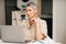 Mature woman in casuals sitting in the living room looking at laptop screen and holding spectacles