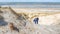 Mature woman climbing a coastal dune among the white sand with her dachshund with the beach and sea in the background