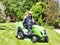 Mature woman driving a tractor lawn mower in garden with flowers