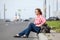 Mature woman in eyeglasses sitting on road curb and hitchhiking, waiting the car