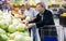 Mature woman with glasses picks a head of cabbage in vegetable section of supermarket