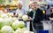 mature woman with glasses picks a head of cabbage in vegetable section of supermarket