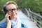 Mature woman with glasses waiting your call. Dressed in a denim jacket, a senior Caucasian lady is standing by river bridge