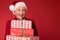 Mature woman in red santa claus hat holding stack of gift boxes, on red studio background. Christmas shopping, copy