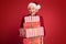 mature woman in red santa claus hat holding stack of gift boxes, on red studio background. Christmas shopping, copy