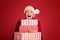 Mature woman in red santa claus hat holding stack of gift boxes, on red studio background. Christmas shopping, copy