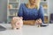 Mature woman sitting at table with piggy bank, using calculator and planning her budget