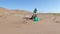 Mature woman in white cap and green gloves digging up a large nylon on the beach and putting it in a garbage bag.