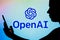 May 2, 2022, Brazil. In this photo illustration, the OpenAI logo is seen in the background of a silhouetted woman holding a mobile