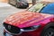 Mazda car grille with chrome logo and front hood with reflections of residential buildings on a sunny day. Red car parked on the
