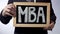 MBA written on blackboard, business person holding sign, business education