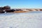 Meadow covered with snow, line of yellow dry reeds on other hill, trees on horizon, winter landscape, blue sky