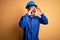 Mechanic man with beard wearing blue uniform and safety helmet over yellow background Shouting angry out loud with hands over