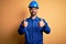 Mechanic man with beard wearing blue uniform and safety helmet over yellow background success sign doing positive gesture with