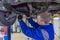 Mechanic working under a car at the garage. Technician wearing blue coverall and using a wrench. Car is on a hydraulic lift