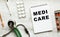 MEDI CARE is written in a notebook on a white table next to pills and a stethoscope