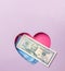 A medical blue mask and a fifty-dollar bill are in a pink heart on a purple background. The minimal concept of money, love and