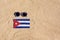 A medical mask in the color of the Cuba flag lies on the sandy beach next to the glasses.