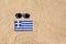 A medical mask in the color of the Greece flag lies on the sandy beach next to the glasses.