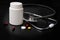 Medical stethoscope lies on a black background around a white empty medicine can, next to pills and tablets, form for the designer