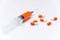 A medical syringe with a orange solution lies on a white background surrounded by pills