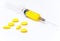 A medical syringe with a yellow solution lies on a white background surrounded by pills