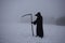 Medieval plague doctor stands on winter field with scythe