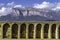 Medieval stone arches aqueduct fortress with the Pyrenees mountain in the background and clouds over blue sky