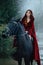 Medieval woman princess in red dress sits astride black steed horse. Girl rider in vintage cloak cape train flies in