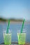 Medium shot of a glass filled to the top green drinking liquid with ice cubes,two straws with blurred ocean background