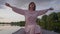 Medium shot of joyful relaxed queer person in pink dress stretching hands sitting in boat on summer sunset on lake