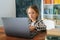 Medium shot of serious primary child school girl sitting at table with laptop and paper book in cozy children room