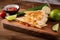 Melty cheese oozing out of a golden-brown quesadilla with fresh salsa on the side and lime wedges on a wooden slicing board