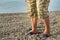 Men& x27;s feet in flip-flops and shorts, a man standing on the beach on a pebble beach, sunrise in the morning