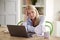 Menopausal Mature Woman Having Hot Flush At Home Cooling Herself With Fan Connected To Laptop