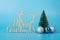 Merry Christmas concept. Photo of small miniature toy evergreen tree three baubles and greeting text isolated over blue color