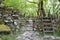 A metal gate through a wall and a stile over it, lead into a rocky path through woodland