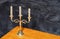 A metal gilded candlestick with three candles stands on a yellow wooden table against a dark gray velor background