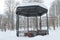 Metal open gazebo with benches in park, covered with snow on cloudy day