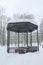 Metal open gazebo with benches in park, covered with snow on cloudy day