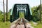 Metal railway bridge with railway tracks and wooden sidewalk running through middle surrounded with utility poles and overgrown fo