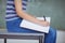 Mid section of schoolgirl sitting on bench and writing on book in classroom