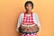 Middle age african american woman wearing baker apron holding homemade cake making fish face with mouth and squinting eyes, crazy