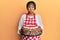 Middle age african american woman wearing baker apron holding homemade cake puffing cheeks with funny face