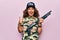 Middle age beautiful hunter woman wearing camouflage t-shirt and usa cap holding shotgun pointing thumb up to the side smiling