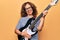 Middle age beautiful musician woman playing electric guitar over isolated yellow background looking positive and happy standing