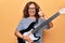 Middle age beautiful musician woman playing electric guitar over isolated yellow background smiling happy and positive, thumb up